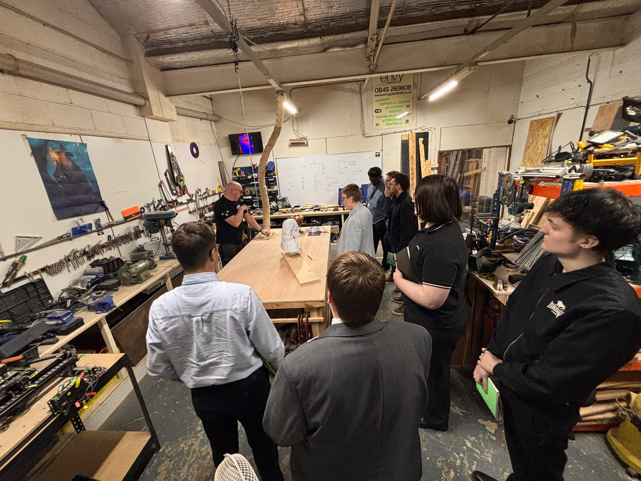 A wide view of Harrison College visitors gathered around a central workbench in the Enigma workshop, with scenery props, tools and materials visible around them.