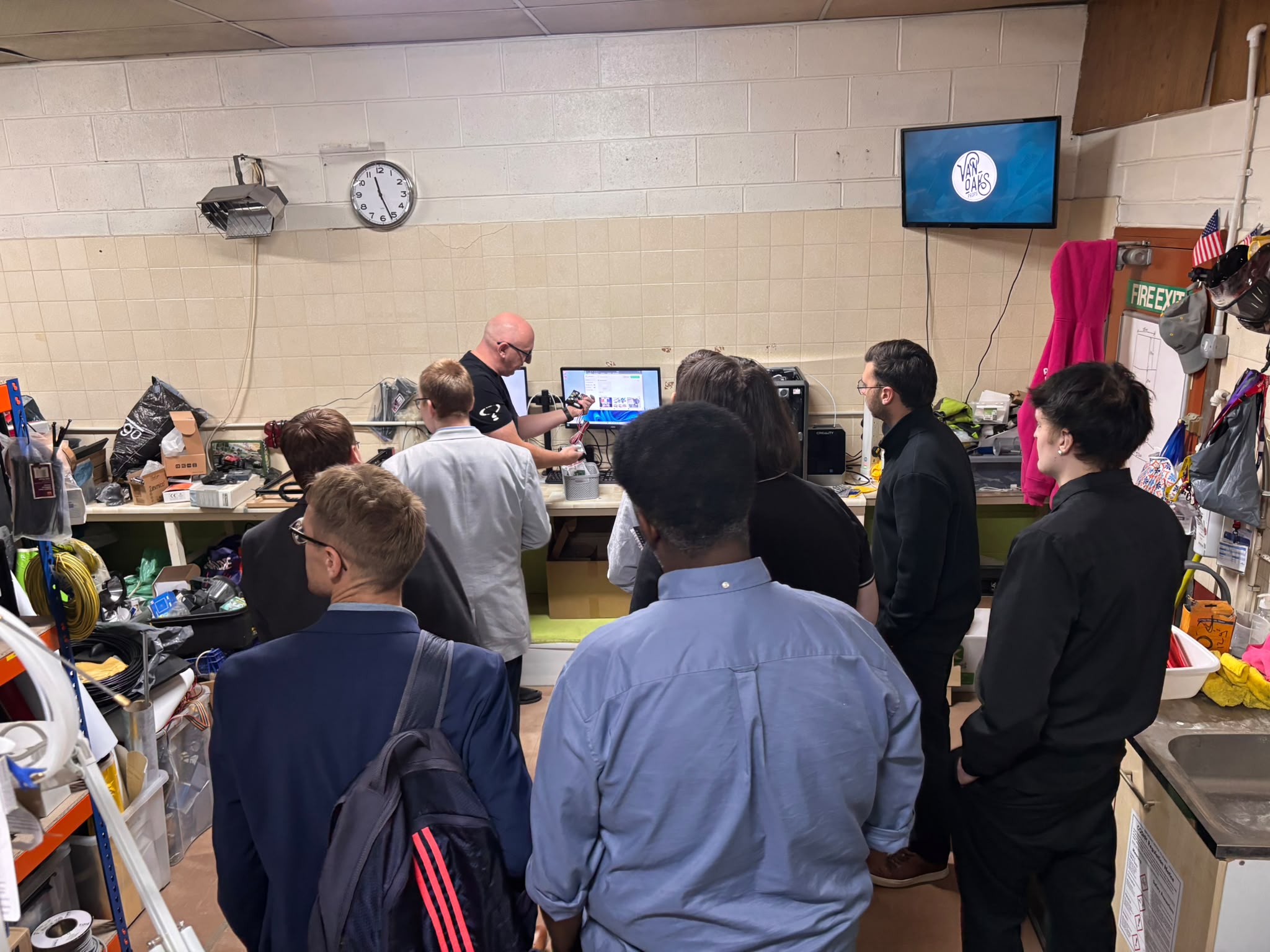 Harrison College visitors watch an Enigma team member demonstrate components at a workbench with computer screens, cables, tools and storage trays.