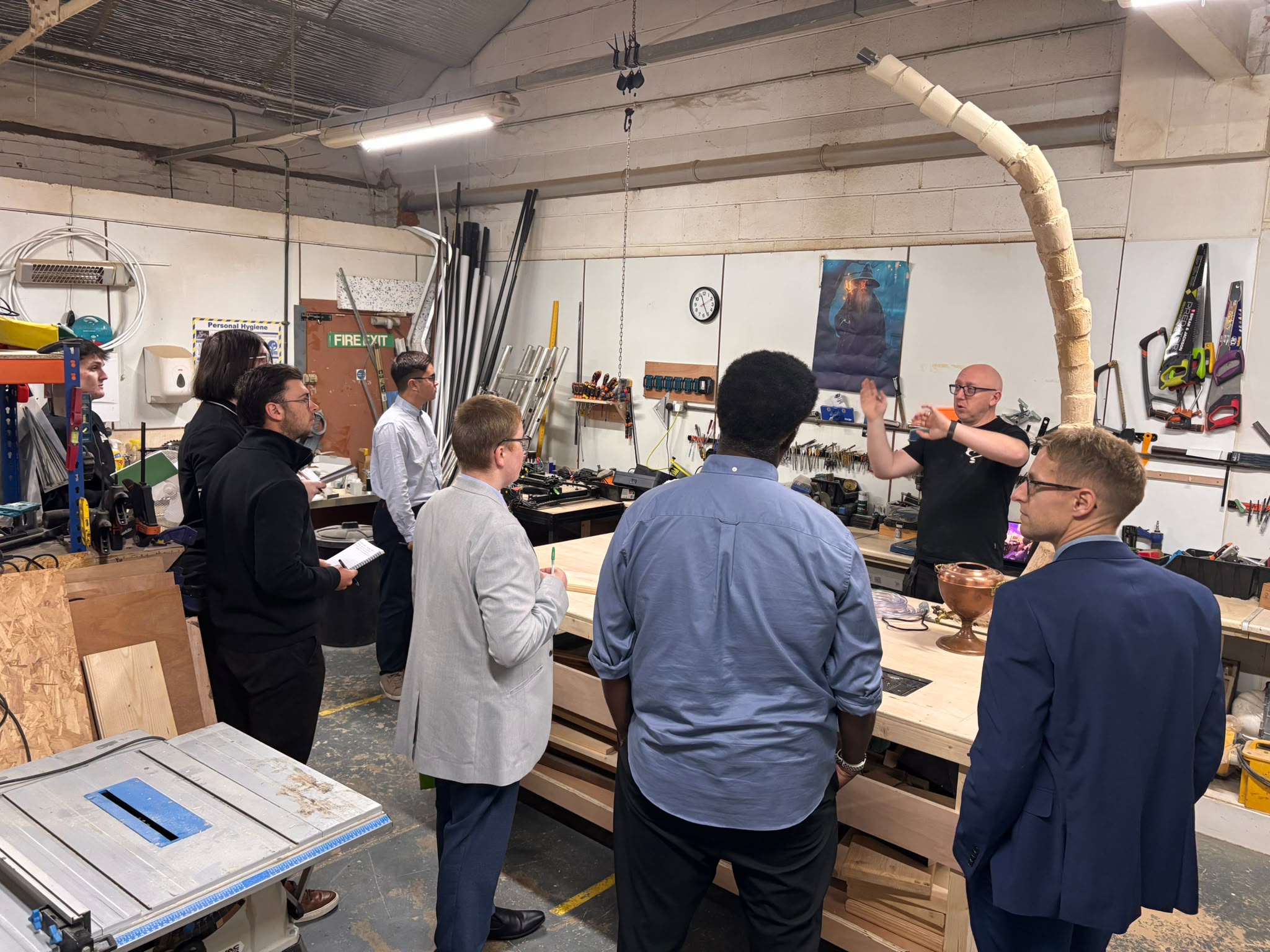 Harrison College visitors gather around a wooden workbench while an Enigma team member explains the making process, with tools and an unfinished tree prop nearby.