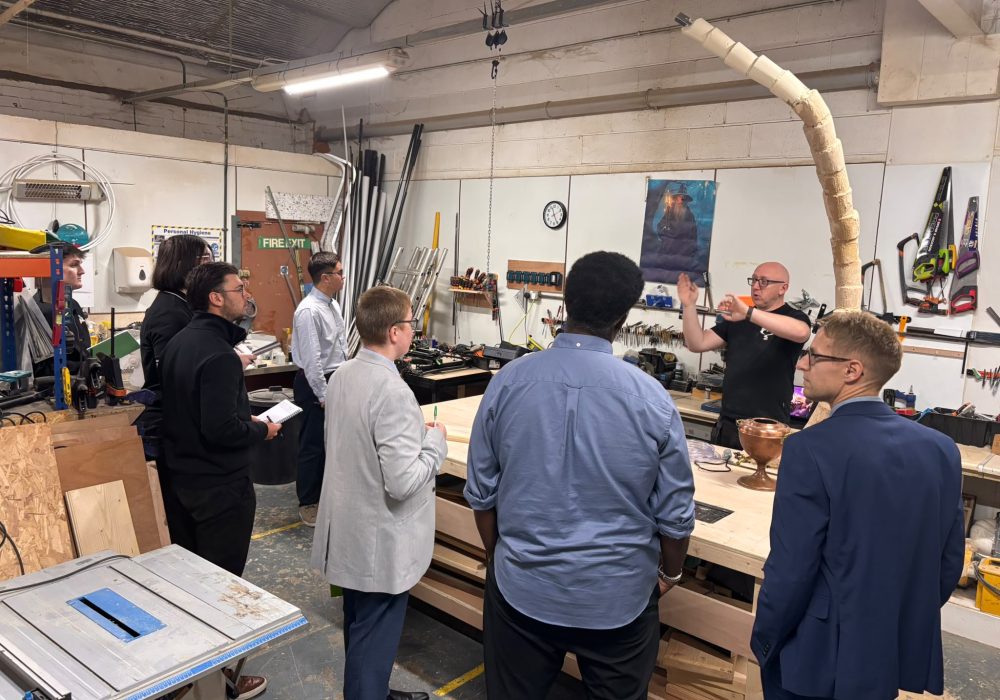 Harrison College visitors gather around a wooden workbench while an Enigma team member explains the making process, with tools and an unfinished tree prop nearby.